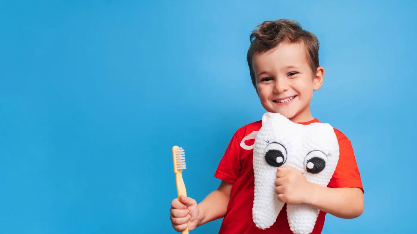 A smiling boy holds a toothbrush and a plush toy tooth against a blue background.