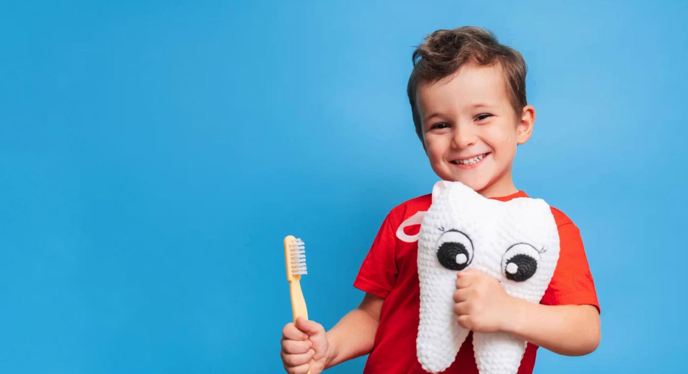 A smiling boy holds a toothbrush and a plush toy tooth against a blue background.