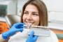 A gloved hand holds a tooth shade guide next to a smiling woman's teeth for color matching.