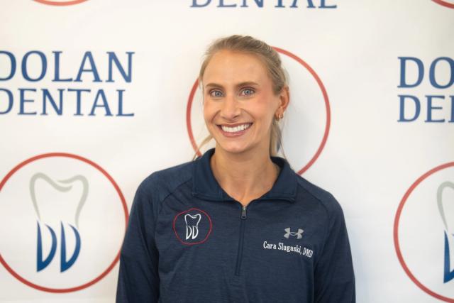 Smiling Dr. Cara Slugaski in a navy shirt with a tooth logo, standing against a Dolan Dental backdrop.