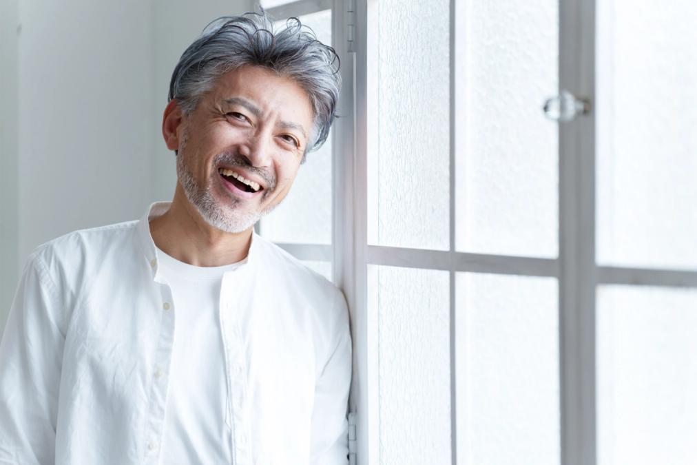 a man with gray hair and a beard is leaning against a window and smiling .