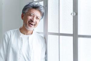 a man with gray hair and a beard is leaning against a window and smiling .