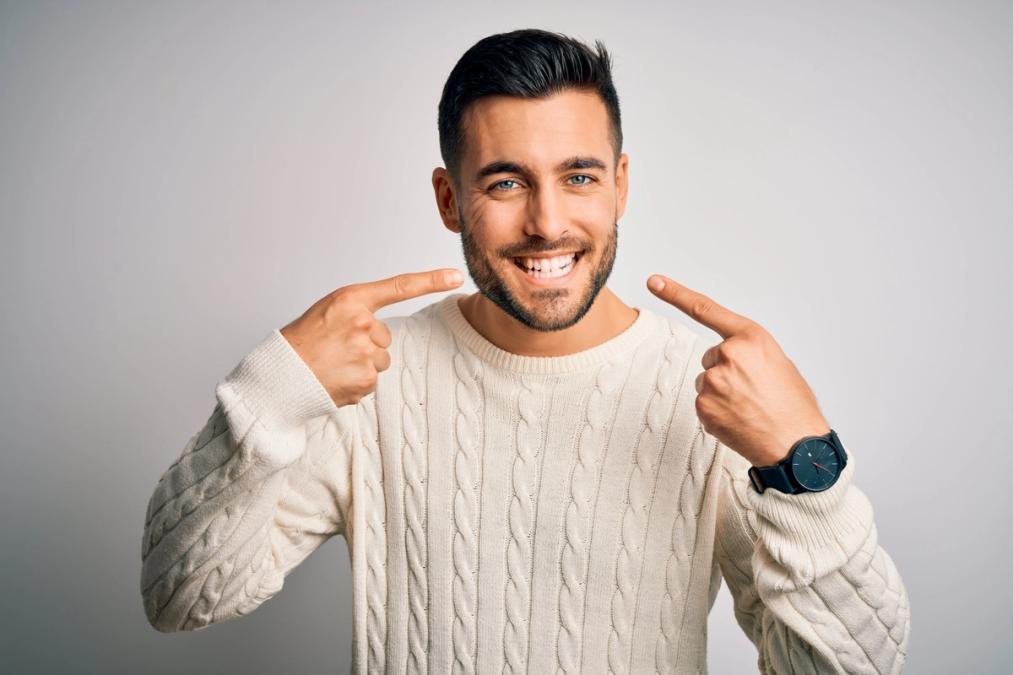 a young man wearing a white sweater is pointing at his teeth and smiling .