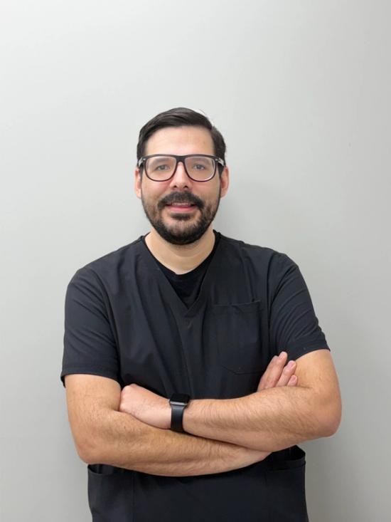 A smiling man in black scrubs and glasses with arms crossed.