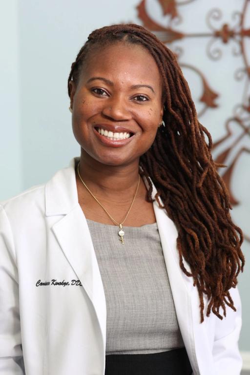 Portrait of a smiling Black woman with long dreadlocks, wearing a white lab coat embroidered with "Canissa Kwabaye, DDS".