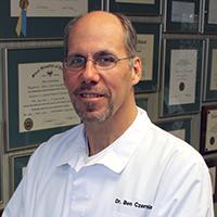 a man wearing glasses and a white coat is standing in front of a wall of framed pictures .