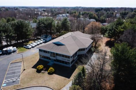 Aerial view of a two-story commercial building with a dark roof, a parking lot with white utility vehicles, and surrounding trees.