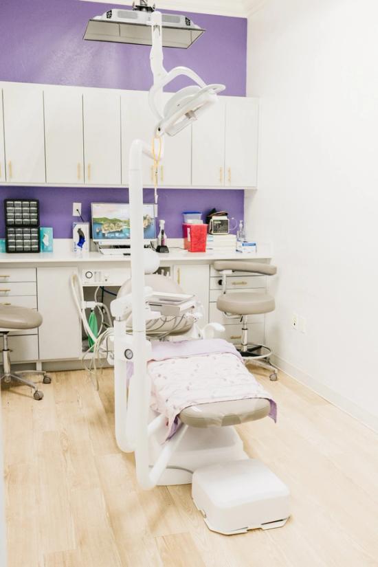 A dental operatory with a purple-blanketed dental chair, overhead light, and a counter filled with equipment set against a purple accent wall.