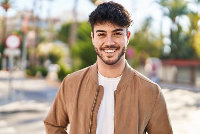A smiling young man with dark hair and a beard wearing a brown jacket.