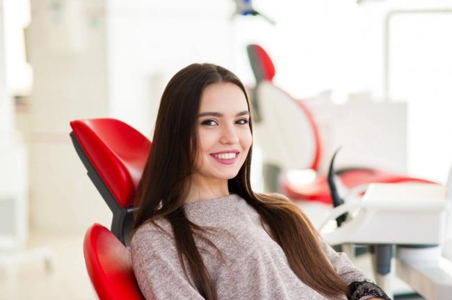 A smiling young woman with long dark hair sits in a red dental chair.