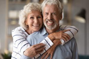 Smiling older woman hugging an older man from behind, both with white hair.