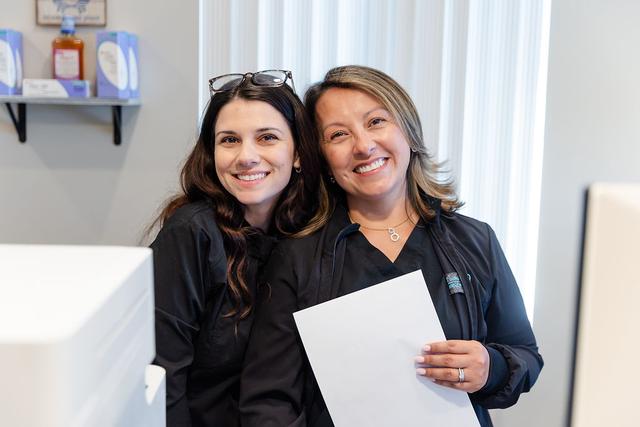 two women are standing next to each other in a room and smiling . one of the women is holding a piece of paper .