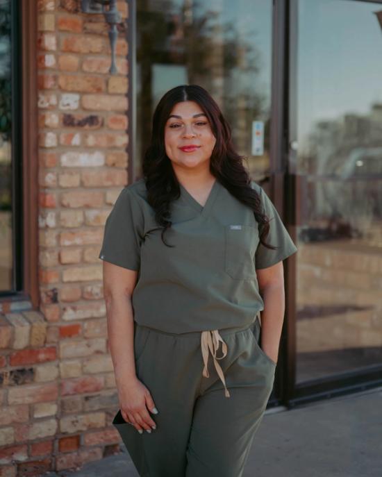 A smiling woman in olive green scrubs stands outdoors.