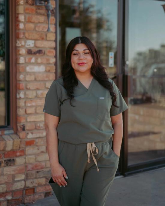 A smiling woman in olive green scrubs stands outdoors.