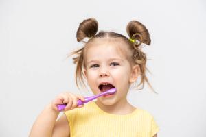 A young girl with pigtails brushes her teeth with a purple toothbrush.