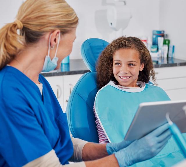 A dental professional shows a tablet to a smiling young girl in a dental chair.