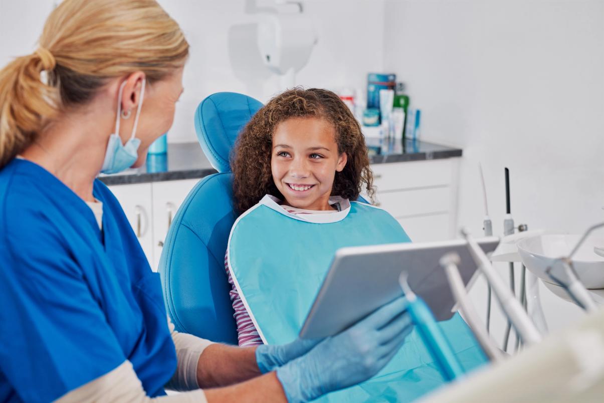 A dental professional holding a tablet speaks to a smiling young girl in a dental chair.