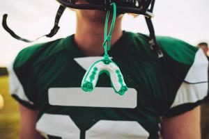 A football player in a green jersey and helmet with a white and green mouthguard hanging from the helmet.
