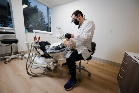 A dentist wearing a white coat, mask, and loupes works on an older patient reclined in a modern dental chair.