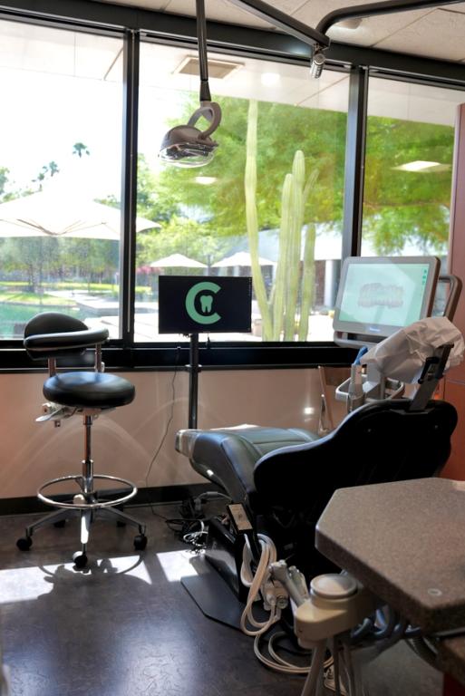 A bright, modern dental operatory with a black examination chair, monitors displaying dental imaging and a logo, and a large window looking out to a sunny desert landscape with cacti.