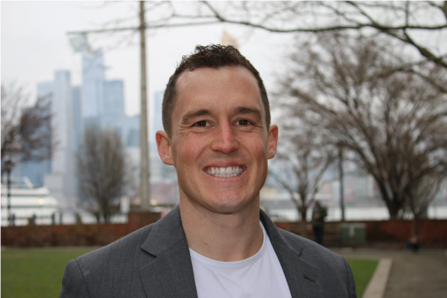 a man in a suit and white shirt smiles in front of a city skyline