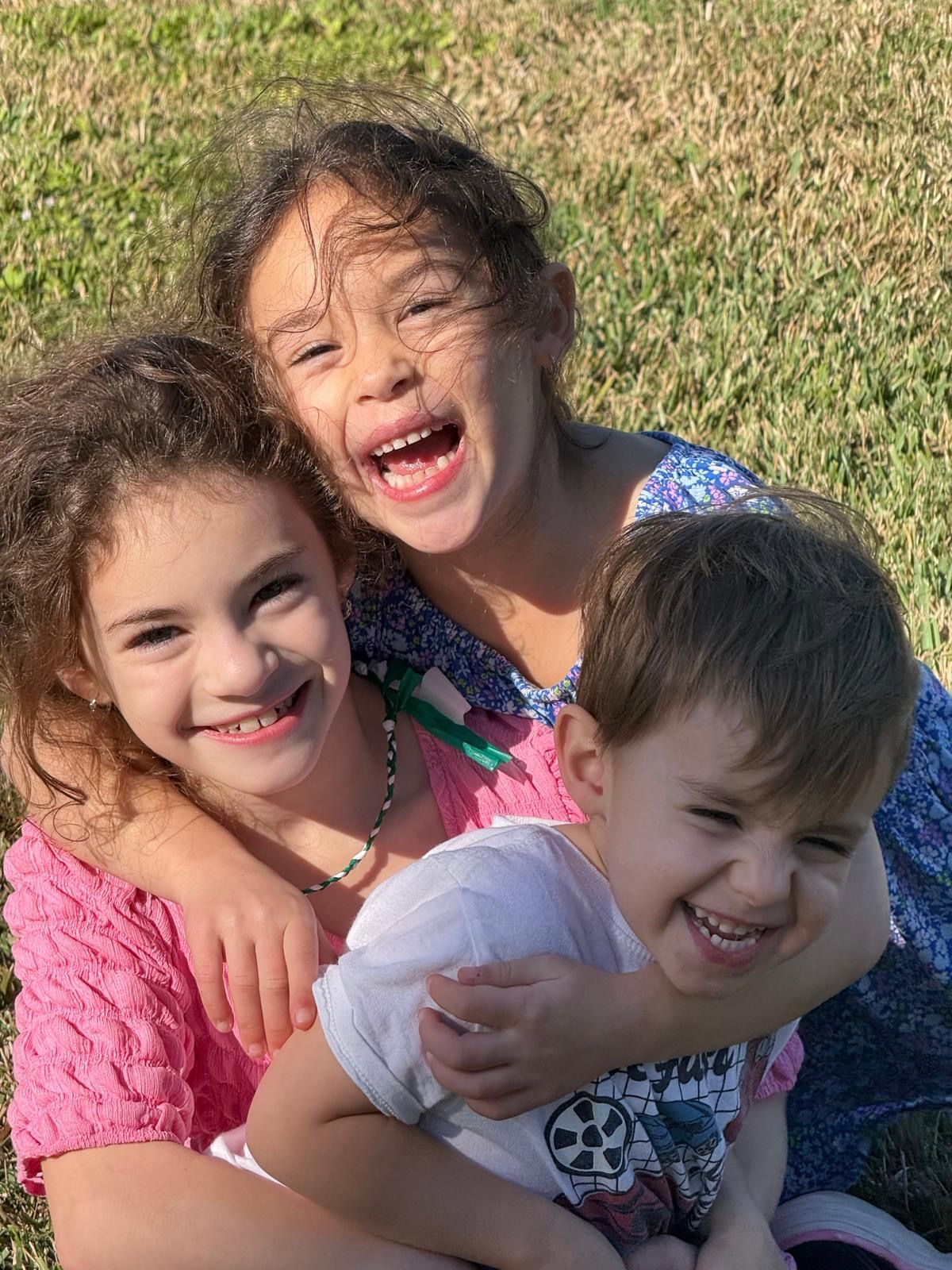 Three young children, two girls and a boy, laughing joyfully together on grass.