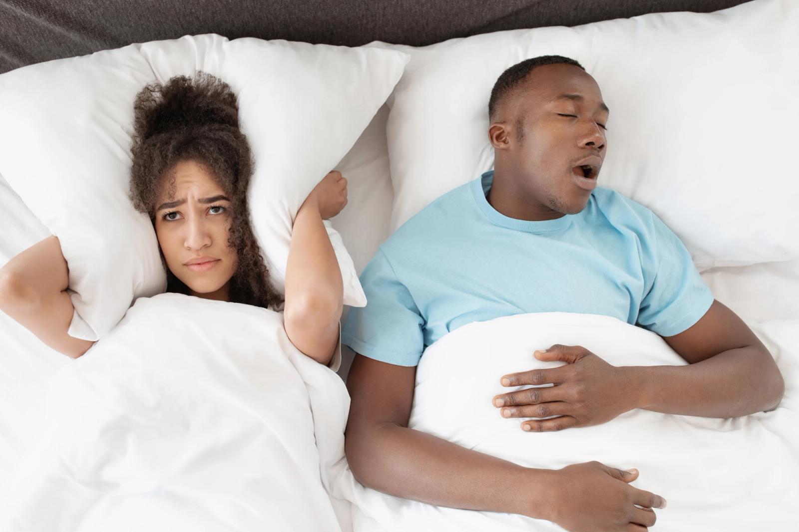 A woman with a pillow over her ears looks annoyed in bed next to a snoring man.