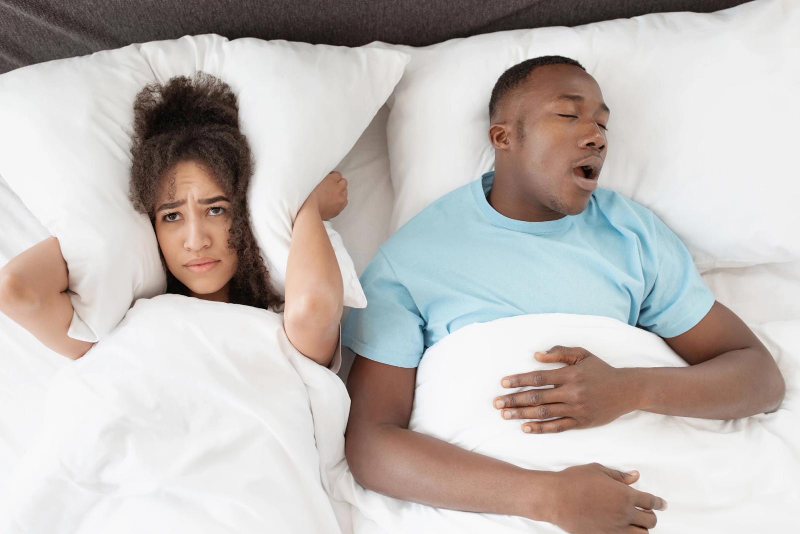 A woman with a pillow over her ears looks annoyed in bed next to a snoring man.