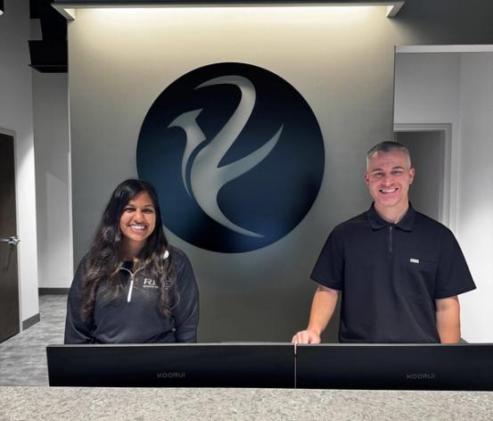 A smiling man and woman stand behind a reception desk with a stylized bird logo on the wall behind them.