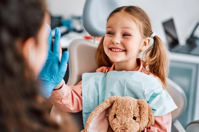 a little girl is sitting in a dental chair holding a teddy bear and giving the dentist a high five .