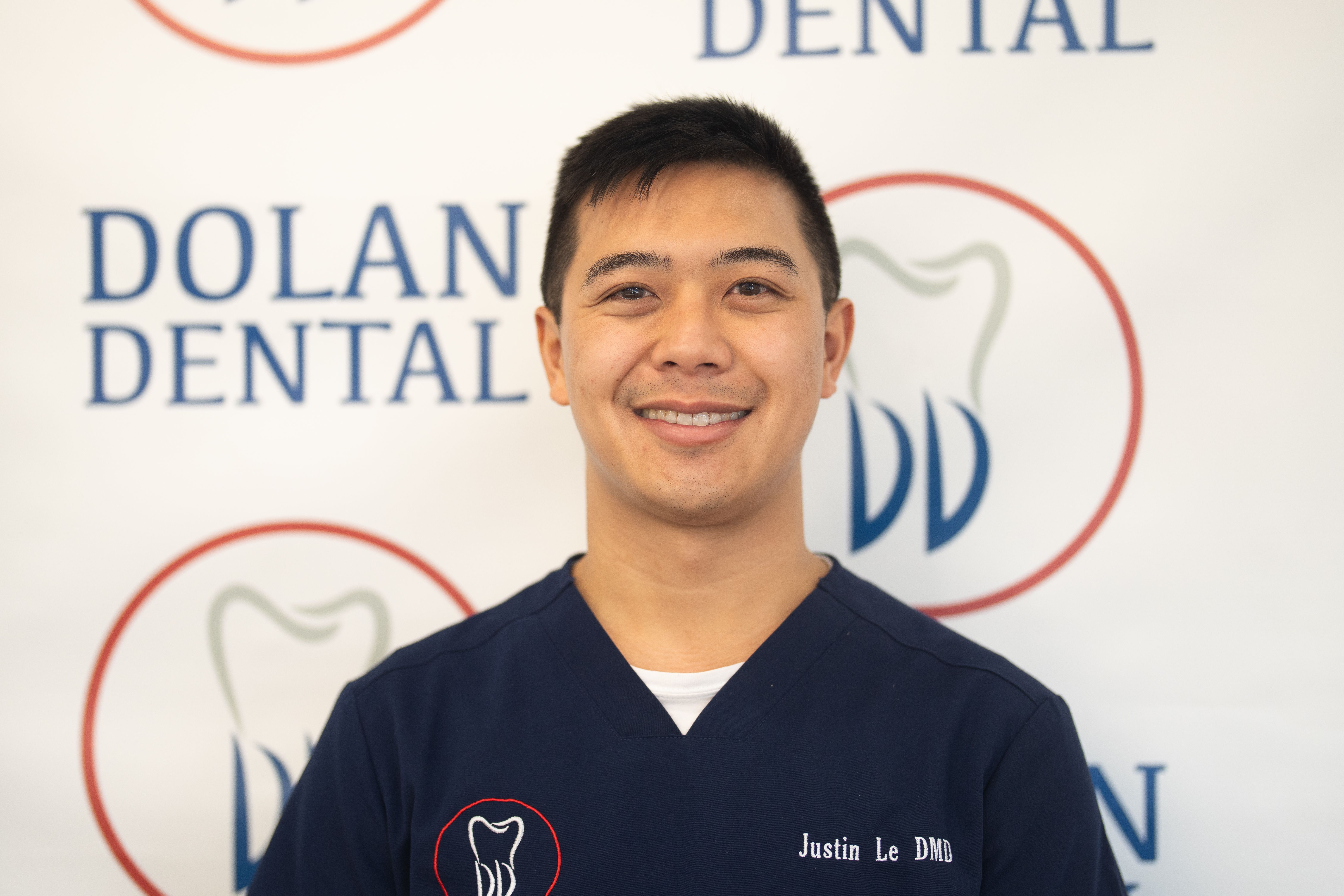 A smiling man in blue scrubs stands in front of a Dolan Dental sign.