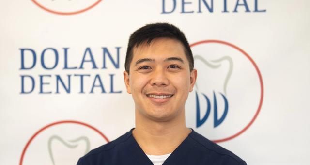 A smiling man in blue scrubs stands in front of a Dolan Dental sign.