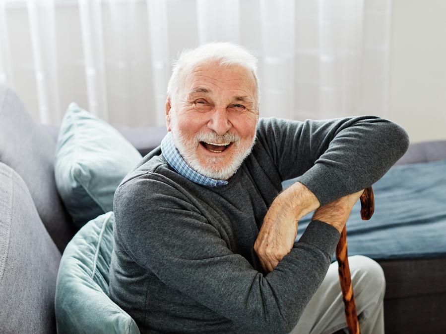 An elderly man with white hair and a beard laughs while sitting on a couch, leaning on a walking cane.
