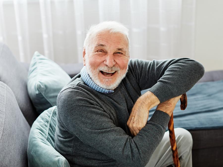 An elderly man with white hair and a beard laughs while sitting on a couch, leaning on a walking cane.