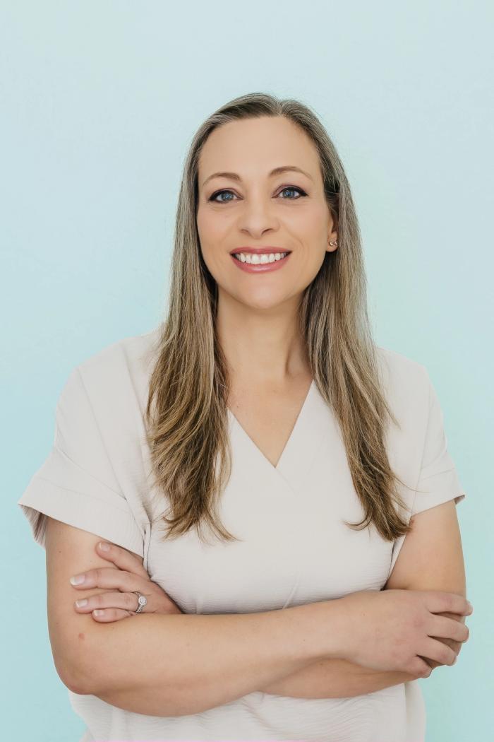 Smiling woman with long hair and arms crossed against a blue background.