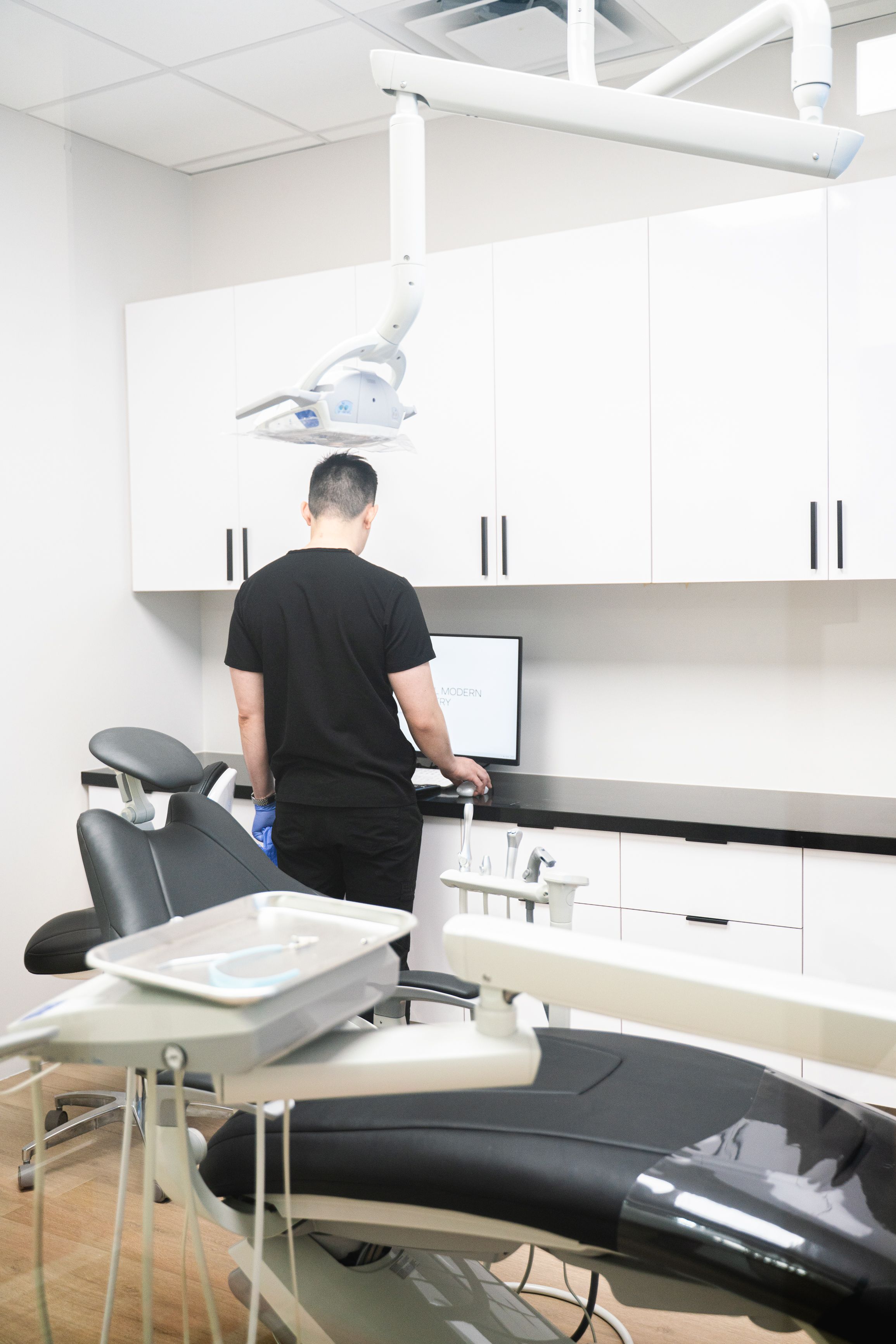 A person in black scrubs stands at a computer in a modern dental operatory, with a dental chair and equipment in the foreground.