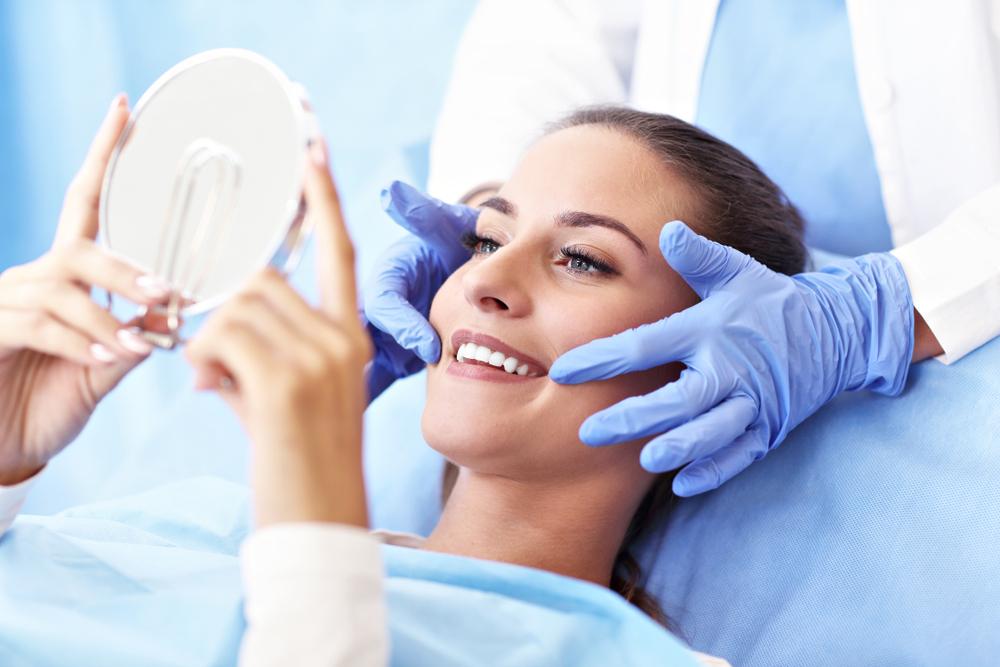 Woman in a dental chair smiling and looking in a mirror, while a gloved hand holds her cheek.