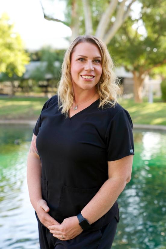A smiling woman with blonde hair wearing black scrubs stands outdoors by a pond.