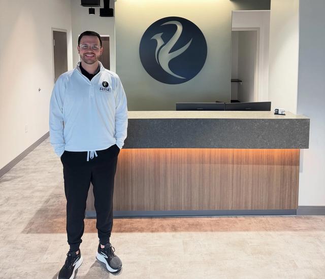 A smiling man in a white shirt stands in a modern reception area with a lit reception desk and a large circular company logo on the wall.