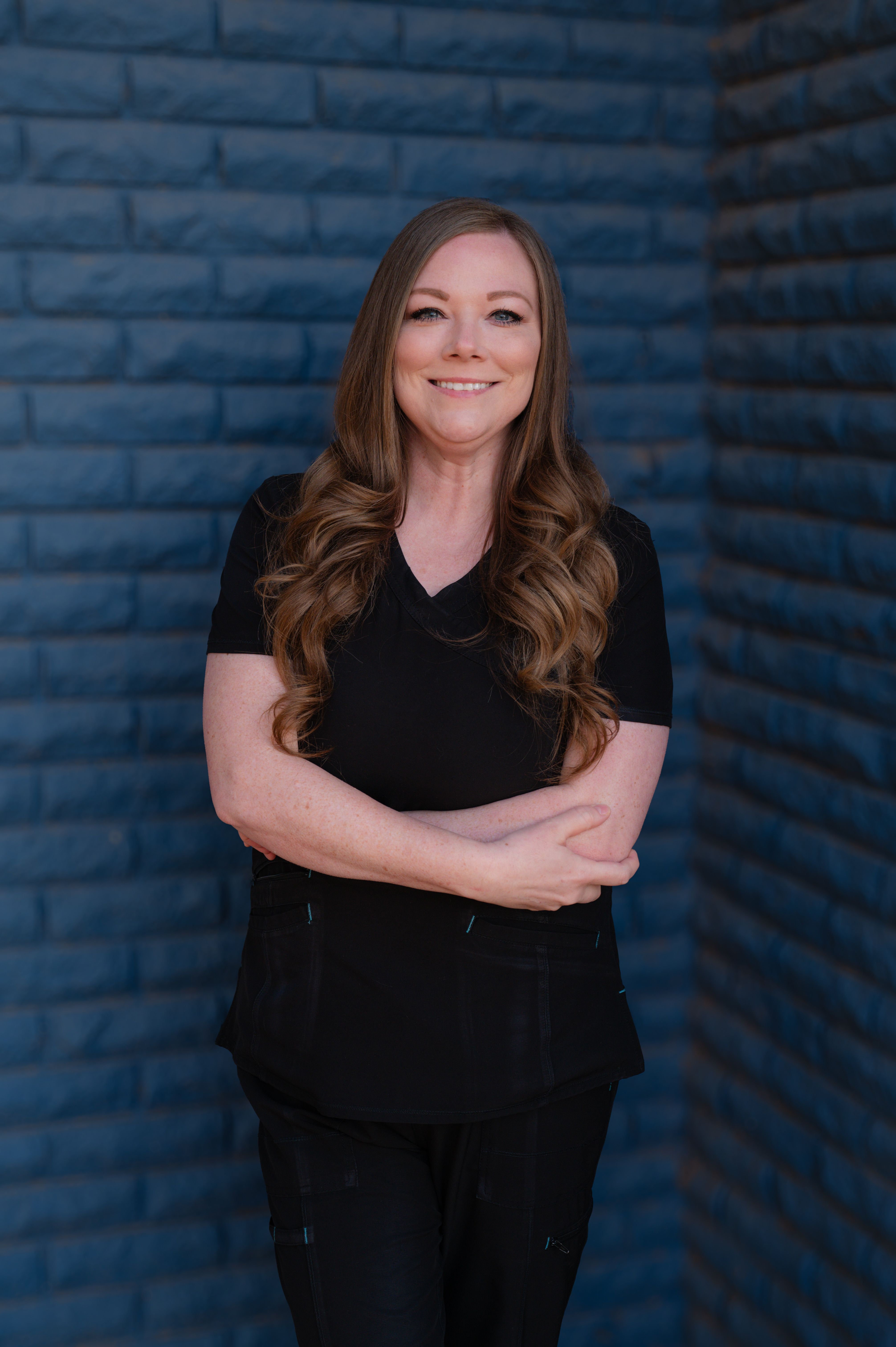 a woman in a black scrub top stands with her arms crossed in front of a blue brick wall