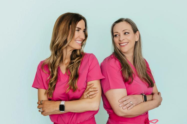 Two smiling women in pink scrubs with crossed arms, looking at each other.