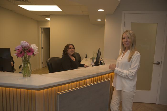 Two women in a modern reception area, one standing by the counter and one seated behind it.
