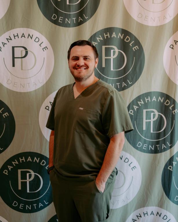 A smiling man in olive green scrubs stands against a Panhandle Dental logo backdrop.