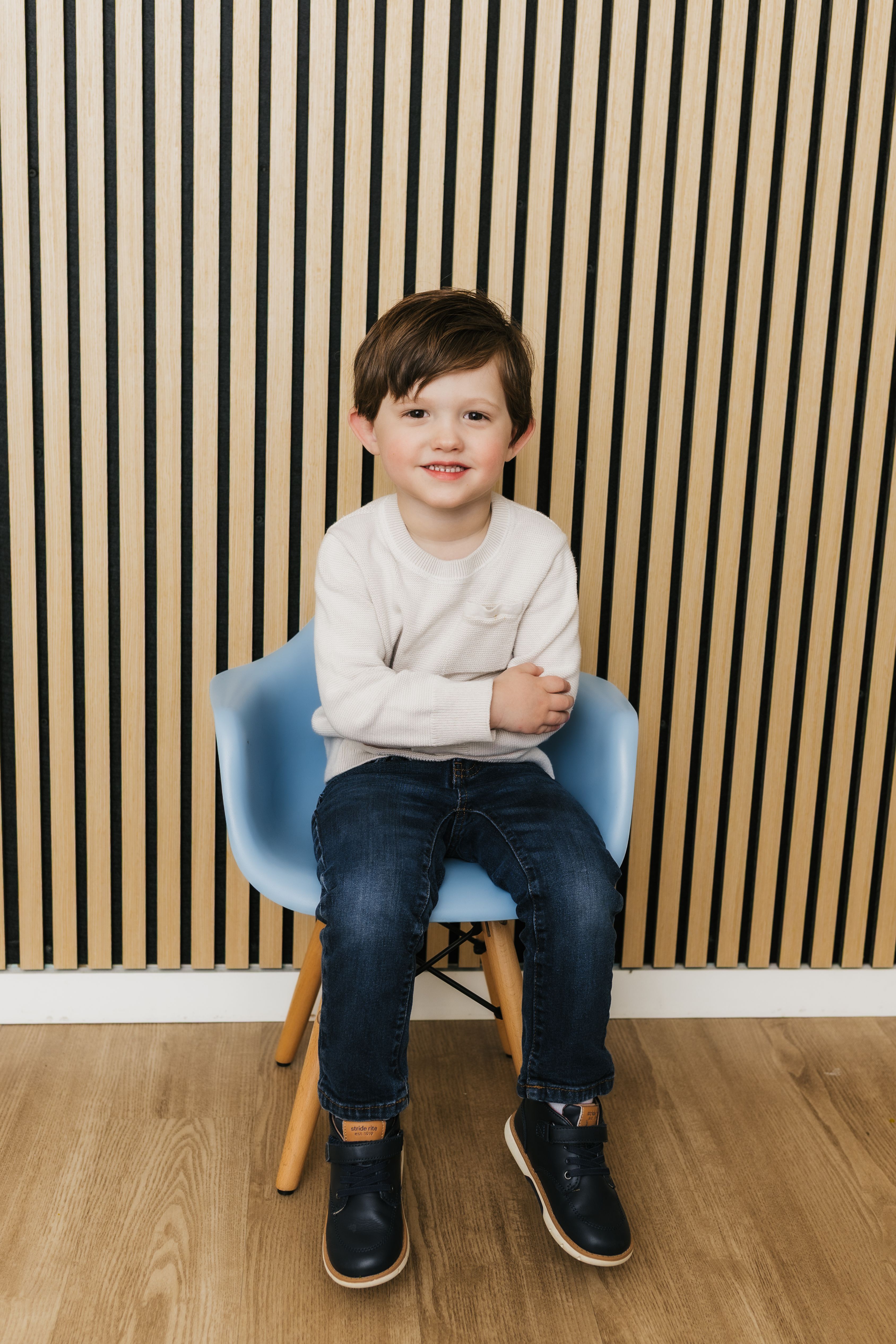 A young boy sits in a blue chair with his arms crossed, smiling at the camera, against a vertically striped wooden wall.