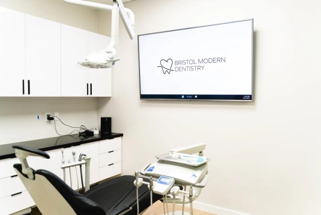 A modern dental examination room with a black dental chair, white cabinets, and a TV displaying the Bristol Modern Dentistry logo.
