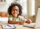 Smiling child with curly hair and glasses writing at a desk with a laptop.