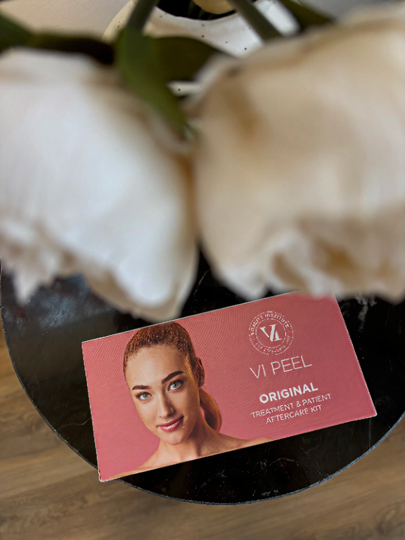 A pink box for VI PEEL Original Treatment & Patient Aftercare Kit on a table, partially obscured by blurred white flowers.