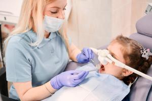 A dentist examines a child in a dental chair who is wearing a nasal mask for sedation.