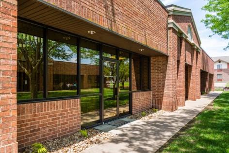 Exterior of a red brick building with large reflective windows, a glass door, a sidewalk, and green grass.