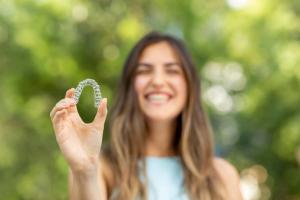 A smiling woman, blurred, holds a clear dental aligner in focus.